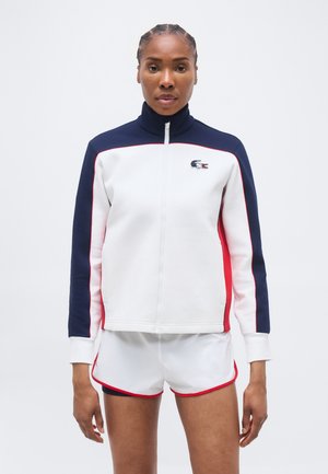 Γυναίκα με πλεξούδες, που φοράει λευκό, ναυτικό και κόκκινο τζάκετ με φερμουάρ και λευκό αθλητικό σορτς με κόκκινη επένδυση, στέκεται μπροστά σε μια απλή φόντο.