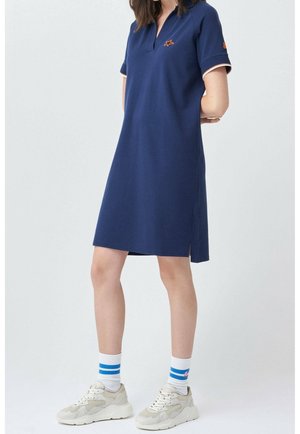 Femme portant une robe polo bleu marine à manches courtes, des baskets blanches et des chaussettes blanches avec deux rayures bleues, debout les mains derrière le dos.