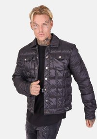 Veste matelassée noire avec fermeture à boutons-pression, texture matelassée, poches poitrine et col court. Portée par-dessus une chemise noire.