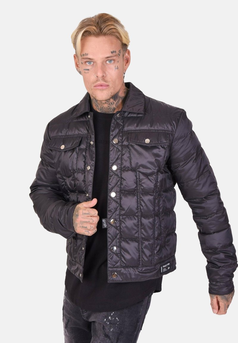 Giacca nera imbottita con chiusura a bottoni a pressione, texture trapuntata, tasche sul petto e colletto corto. Indossata sopra una camicia nera.