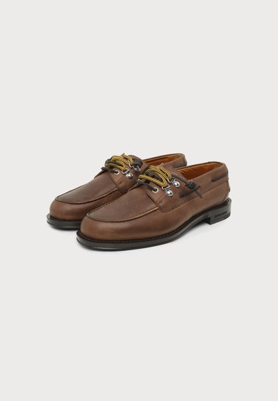 Paire de chaussures bateau en cuir marron avec des lacets jaunes et noirs et des semelles noires sur un fond clair uni.