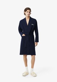 Accappatoio blu navy con cintura, realizzato in tessuto morbido, con un logo sul petto e due tasche frontali, indossato sopra calzature casual.