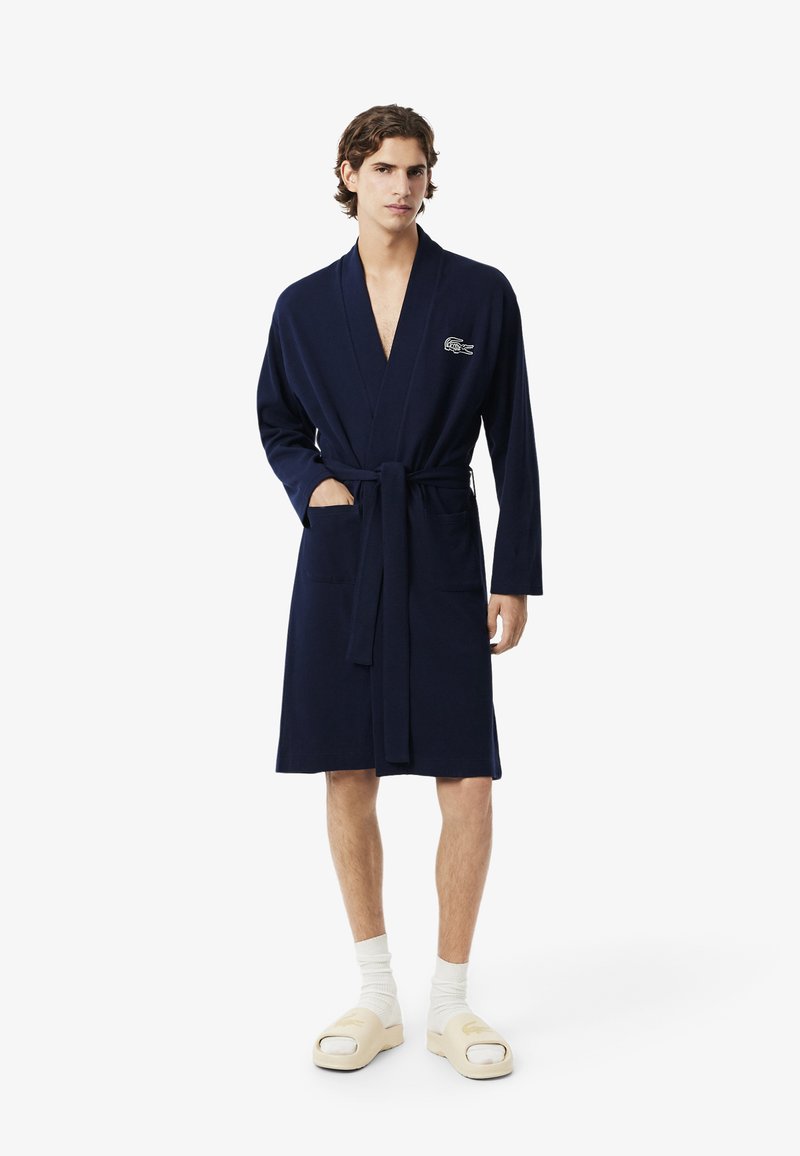 Accappatoio blu navy con cintura, realizzato in tessuto morbido, con un logo sul petto e due tasche frontali, indossato sopra calzature casual.