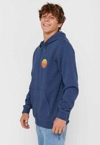 Jonge man die glimlacht, gekleed in een marineblauwe rits hoodie met een klein oranje rond logo en lichtblauwe spijkerbroek, staand tegen een effen achtergrond.
