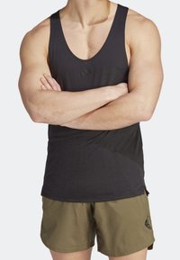 Débardeur de sport noir sans manches avec une texture lisse, arborant un petit logo sur la poitrine, accompagné d'un short vert olive.