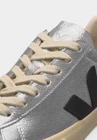 Detailní záběr stříbrné tenisky s krémovými tkaničkami, černým logem "V" na boku a béžovou podrážkou.