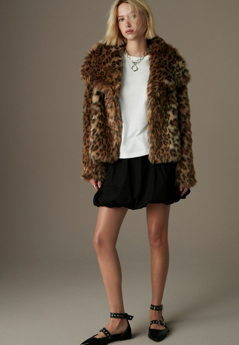Faux fur leopardprint frakke med en voluminøs krave, parret med en hvid T-shirt, sort plisseret nederdel og sorte flade sko med nitter.