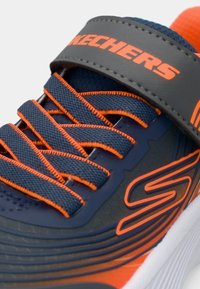 Blizak prikaz mornarsko plavog i narančastog Skechers tenisica s teksturiranim vezicama, remenom, logom i bijelim detaljima na đonu.