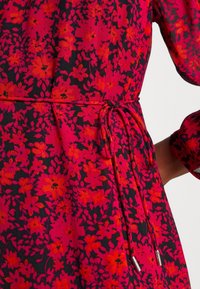 Robe nouée à la taille avec des manches longues présentant un motif floral dense rouge et noir sur un fond rose vif.