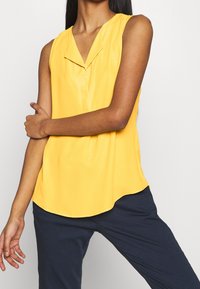 Donna che indossa un blusa gialla senza maniche con dettagli sul collo, abbinata a pantaloni blu scuro, in piedi su uno sfondo chiaro.