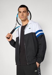 Svart og hvit zip-up jakke med blå striper på ermene, båret av en person som holder en tenniskjøtt. Har et logo på brystet.