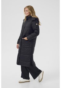 Črn puffer plaš�č z dolgim, quilted dizajnom, ki vključuje kapuco, sprednje žepe in stranski razporek. Nosi se preko temnih širokih hlač in belih superg.