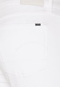 Jeans de mezclilla blanca con un bolsillo trasero que presenta una costura curva y una pequeña etiqueta negra. La tela tiene una textura suave.