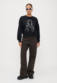 Sweatshirt preto com um design em arte linear branca de uma pessoa, combinado com calças largas castanhas e ténis robustos pretos. Óculos de sol transparentes usados.