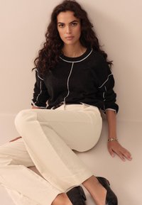 Maglione nero con dettagli bianchi, pantaloni di velluto a coste beige, scarpe con tacco nere e braccialetto d'argento. Soggetto seduto su uno sfondo neutro.