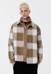 Ung man med kort blont hår som bär en brun och vit rutmönstrad knappjacka över en brun zip-up tröja med händerna i fickorna.