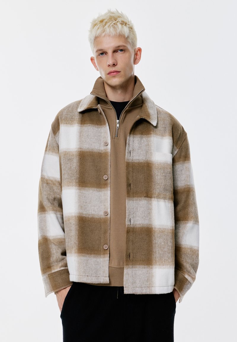 Ung man med kort blont hår som bär en brun och vit rutmönstrad knappjacka över en brun zip-up tröja med händerna i fickorna.