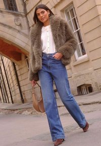 Dirbtinės odos striukė alyvų žalia spalva, balta palaidinė su mygtukais, aukšto liemens mėlynos džinsai, rudi aukštakulniai sandalai ir ruda pečių rankinė.