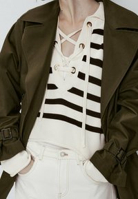 Manteau trench olive sur un haut en tricot blanc et noir à encolure lacée. Jeans blancs visibles avec un détail de bouton.