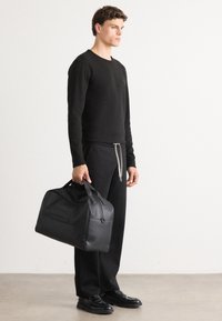 Sort læder duffelbag med dobbelte håndtag, rektangulær form, glat tekstur og synlig lynlåslomme på siden.