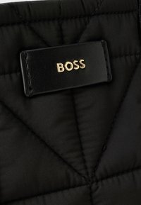 Fekete steppelt anyag, textúrált felülettel, fekete bőrcímkével, rajta arany "BOSS" logóval. Geometrikus varrásminta látható.