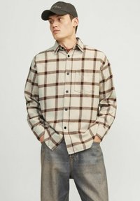 Chemise en flanelle beige à motif carreaux marron, avec un col boutonné, une poche poitrine unique et des manches longues, associée à un jean en denim.
