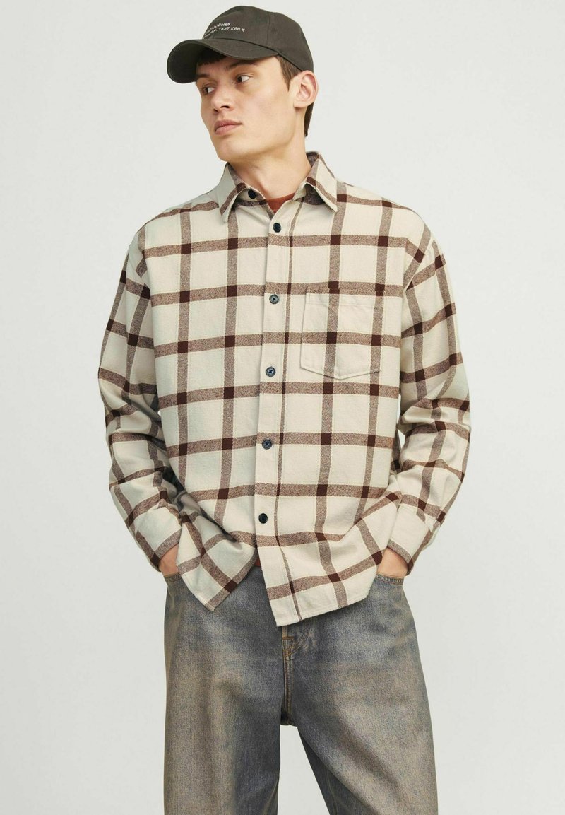 Chemise en flanelle beige à motif carreaux marron, avec un col boutonné, une poche poitrine unique et des manches longues, associée à un jean en denim.