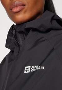 Black windbreaker jacket featuring a textured surface, a high collar, and a zip closure. Decorated with a white "Jack Wolfskin" logo.