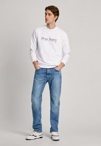 Sudadera blanca con logo azul, vaqueros de un azul claro y zapatillas blancas con acentos negros. Ajuste casual con puños y cuello de canalé.