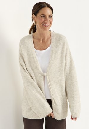 Frau lächelt, trägt eine hellbeige flauschige Strickjacke, die einmal über der Brust über einem weißen Hemd und dunklen Hosen gebunden ist, und steht vor einem einfarbigen Hintergrund.