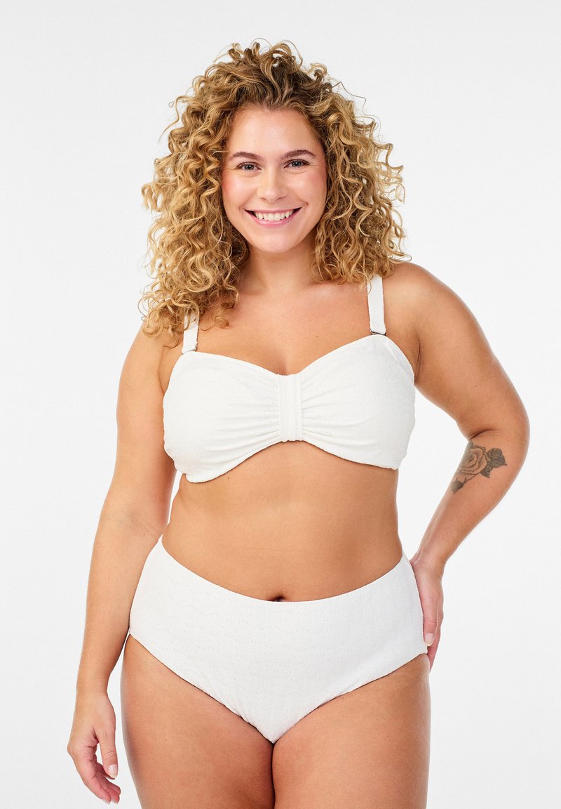 Uśmiechnięta kobieta o kręconych blond włosach, ubrana w biały, teksturowany dwuczęściowy strój kąpielowy, stojąca z jedną ręką na biodrze na białym tle.