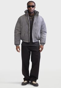 Gray puffy jacket with high collar, paired with black t-shirt and loose-fitting black pants. Accessories include sunglasses and layered gold necklaces.