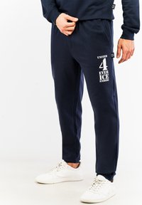 Marineblå joggebukser lavet af bomuldsblanding, med et trykt grafik på låret og et elastisk bånd for komfort. Kombineret med hvide sneakers.
