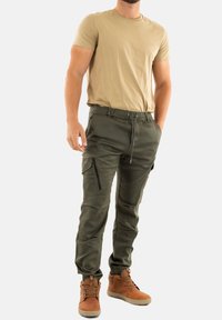 Pantalons cargo vert olive avec poches zippées, taille à cordon de serrage et poignets élastiques ; associés à un t-shirt beige et des bottes à lacets brunes.