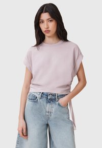 T-shirt cropped di un lilla chiaro con maniche corte arrotolate, dettaglio con cordoncino sul lato, abbinato a jeans blu sbiaditi a vita alta.