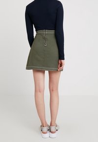 Falda de mezclilla verde con un diseño acampanado, costuras de contraste y un cierre de cremallera en la parte posterior, combinada con una blusa ajustada de manga larga en azul marino y zapatillas blancas.