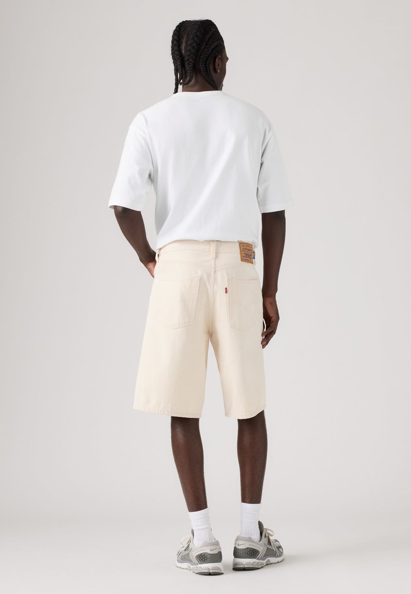 Uomo con capelli intrecciati che indossa una t-shirt bianca oversize, pantaloncini beige fino al ginocchio, calzini bianchi e scarpe da ginnastica grigie, in piedi con le mani dietro la schiena.
