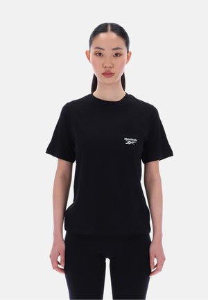 Junge Frau mit dunklem, zurückgebundenem Haar trägt ein schwarzes Reebok-T-Shirt und schwarze Hose, steht vor einem schlichten hellen Hintergrund.