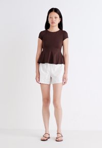 A young woman stands against a white background, wearing a brown peplum top and white shorts, with black sandals and straight hair.