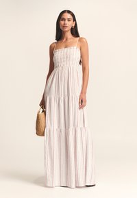 Vestido maxi con corsé fruncido, tela blanca con rayas verticales rojas, tirantes finos, diseño de capas y un bolso tejido con textura.