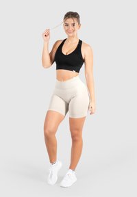 Zwarte geribbelde sportbh met een V-hals, gecombineerd met hooghangende beige shorts. Model staat in witte sportschoenen. Lichte achtergrond.