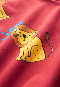 Dessin animé d'un lion aux yeux fermés et au corps tacheté imprimé sur un tissu rouge, montrant une partie d'un autre lion et du texte bleu en arrière-plan.