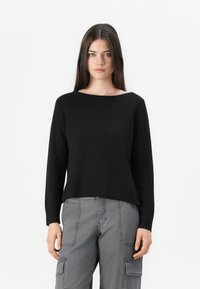 Jeune femme avec des cheveux bruns lisses portant un pull noir à manches longues et un pantalon cargo gris, se tenant contre un fond blanc.