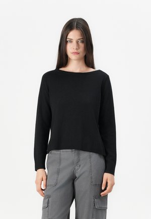 Jeune femme avec des cheveux bruns lisses portant un pull noir à manches longues et un pantalon cargo gris, se tenant contre un fond blanc.