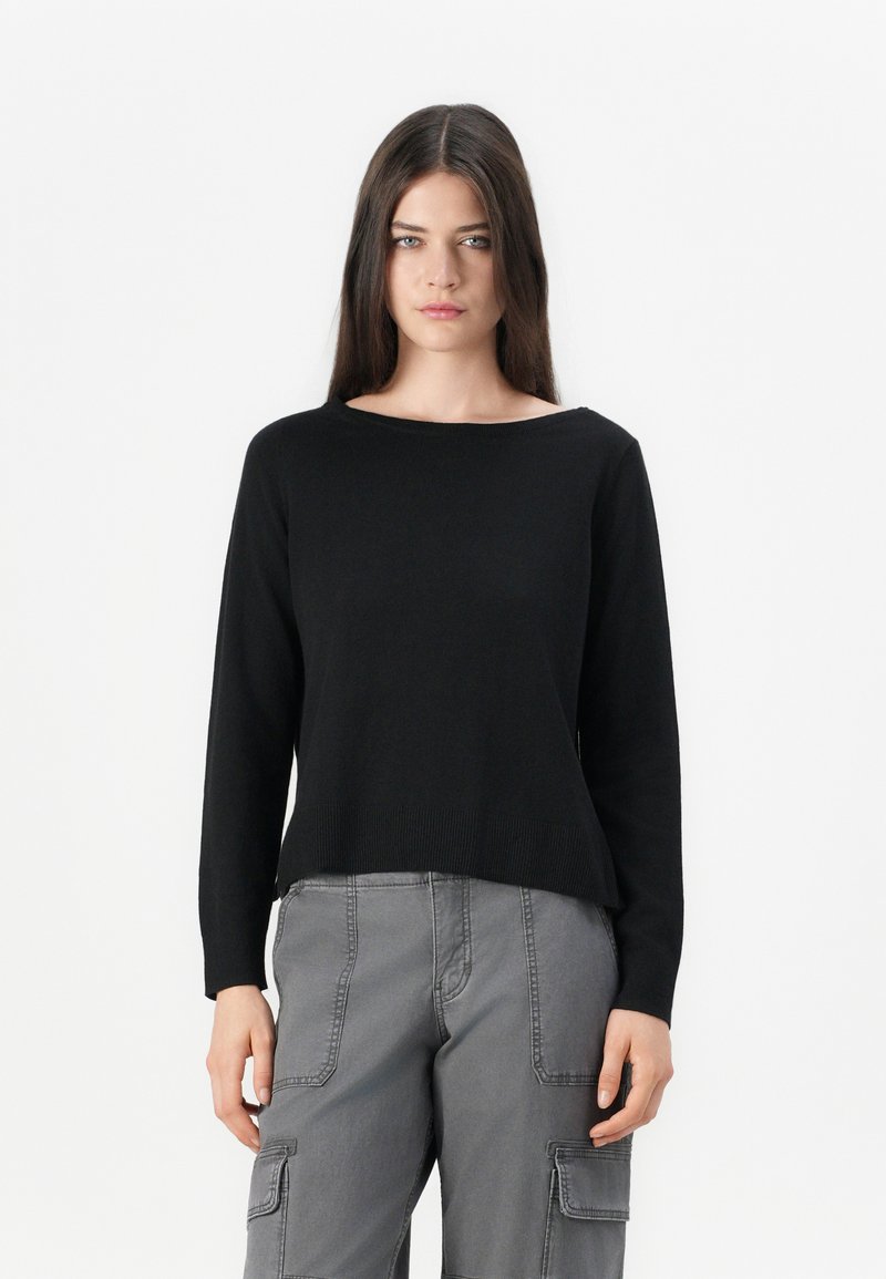 Jeune femme avec des cheveux bruns lisses portant un pull noir à manches longues et un pantalon cargo gris, se tenant contre un fond blanc.