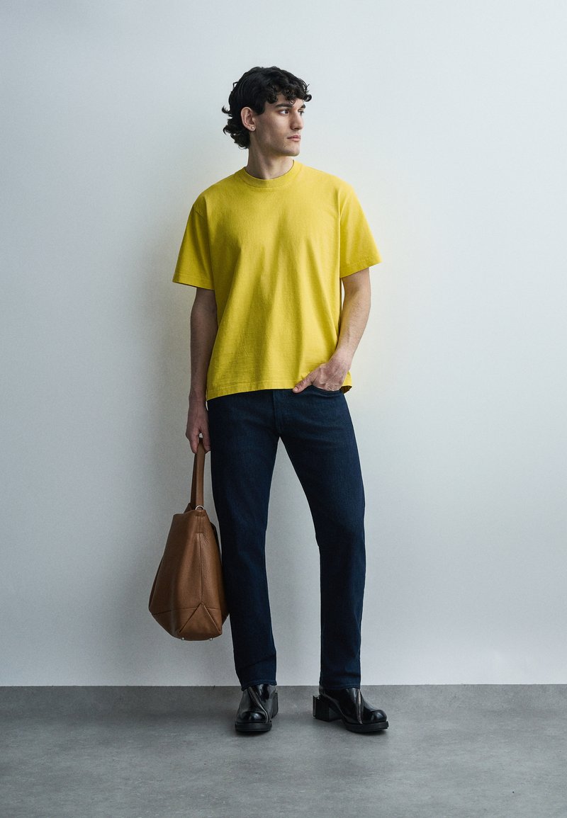 Giovane uomo con una t-shirt gialla, jeans scuri, scarpe nere, che tiene una borsa di pelle marrone, in piedi davanti a una parete grigia chiara uniforme.