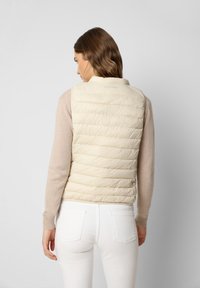 Helles beiges Puffer-Weste mit horizontalen Steppnähten, hohem Kragen und Branding am Hals, getragen über einem beigen Pullover und weißen Hosen.