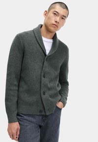 Grå strikket cardigan med sjalkrave, knaplukning og ribbet struktur, parret med en hvid T-shirt og mørke jeans.