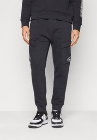 Svarta sweatpants med cargofickor, elastisk midja och präglad logodetalj. Tyget verkar mjukt med en klassisk passform. Matchas med svarta och vita sneakers.