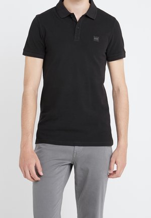 Mand iført sort kortærmet polo med tre knapper og grå bukser, stående med armene afslappet ned langs siderne.